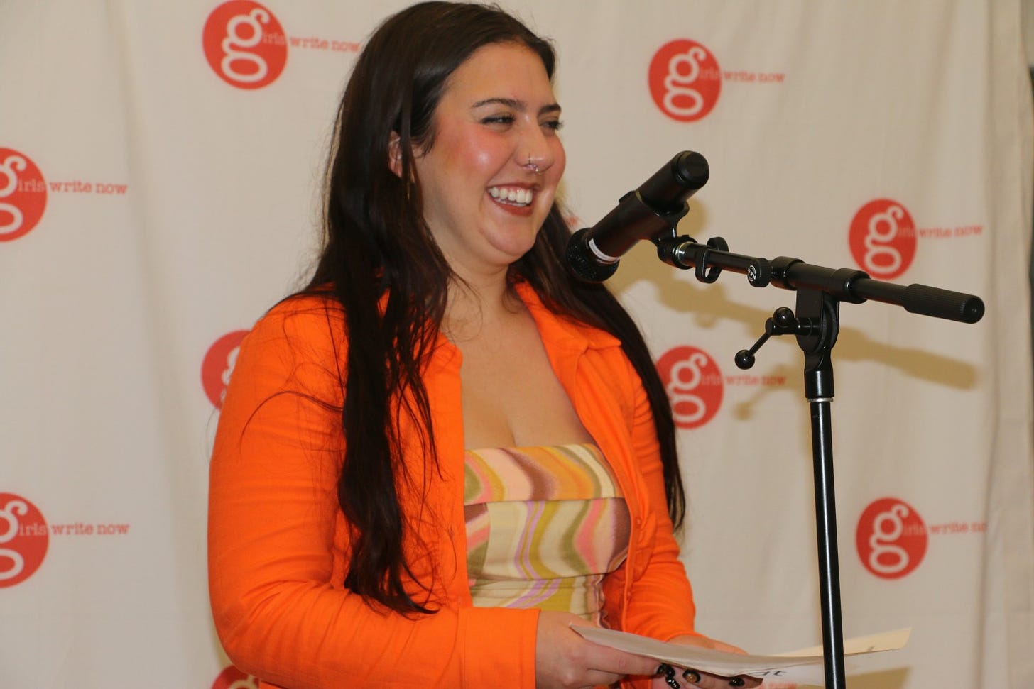 Subject Annabel in an orange jacket and multi-color dress at a mic