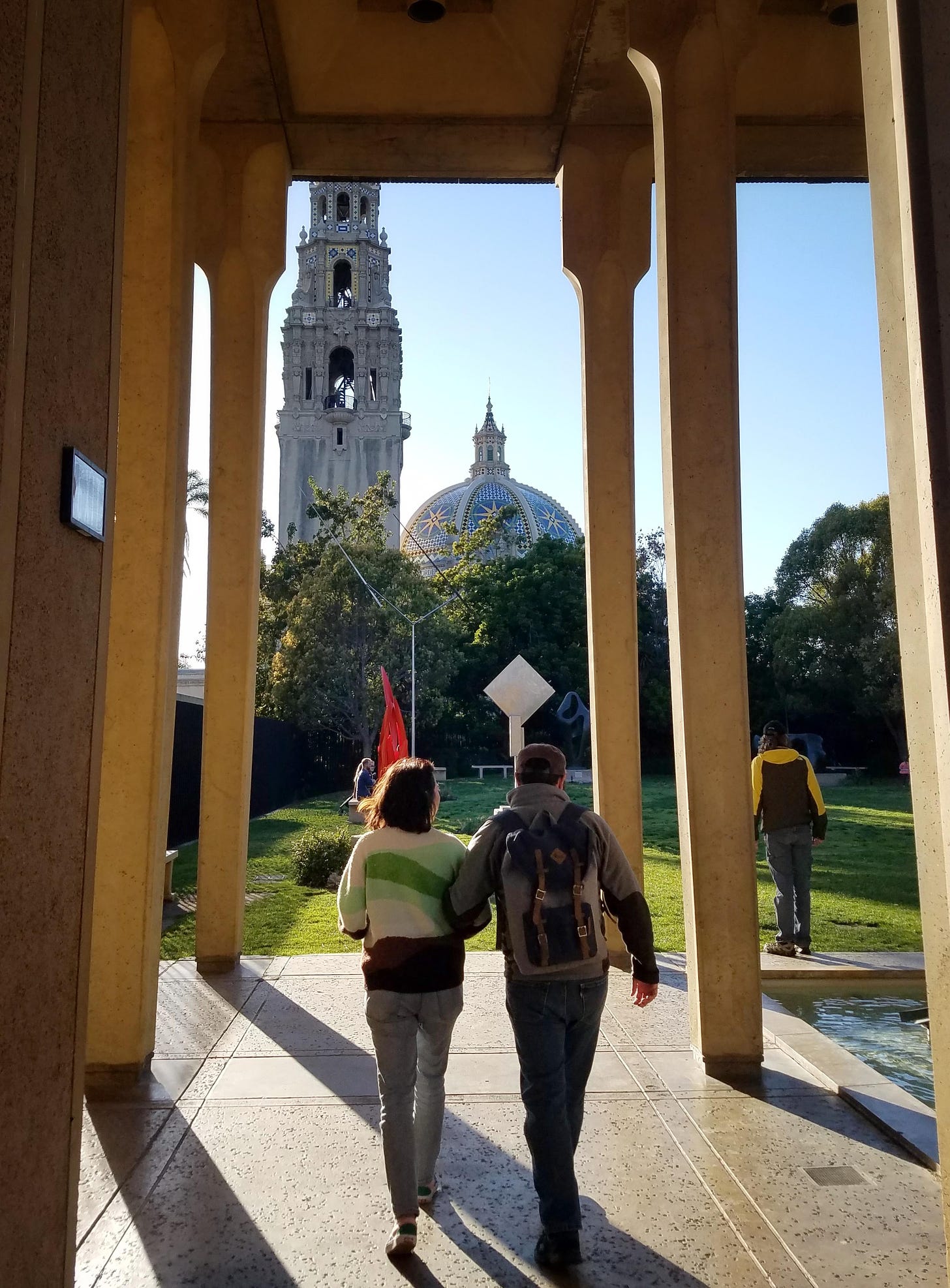 Two people walk arm-in-arm through a corridor leading to a sculpture garden on a lawn, with a domed building and tall bell tower in the background. The person on the left is a petite woman with dark hair and a striped green sweater and jeans; on the right is a man wearing a brown cap, backpack, grey fleece, and jeans. They're walking away from the camera