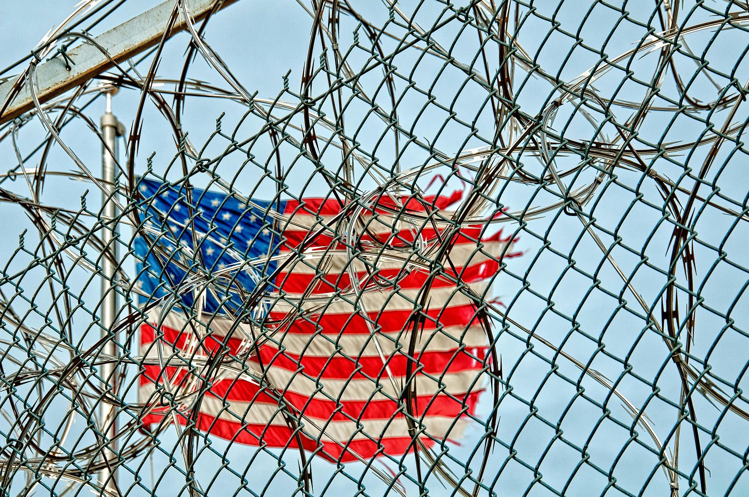 A tattered USA flag flies behind a chain-link fence and barbed wire