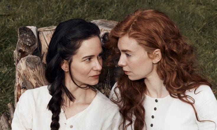 Tallie e Abigail deitadas na cama e olhando. Ambas vestem branco