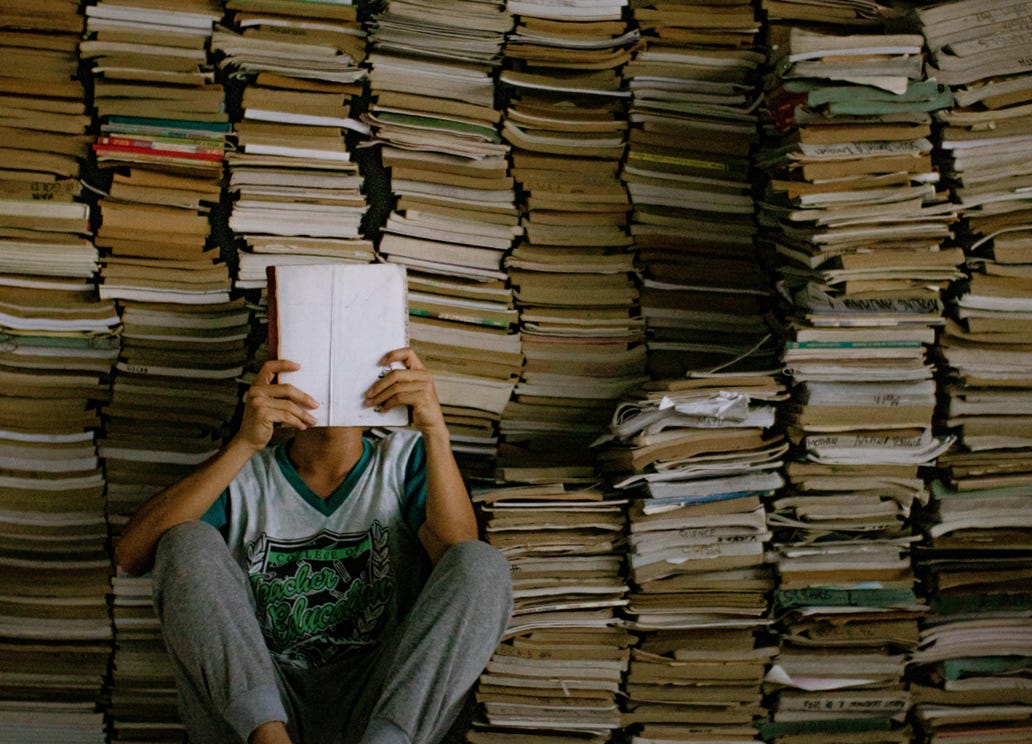 man in gray sweatpants with a book covering his face leans against a wall stacked with books