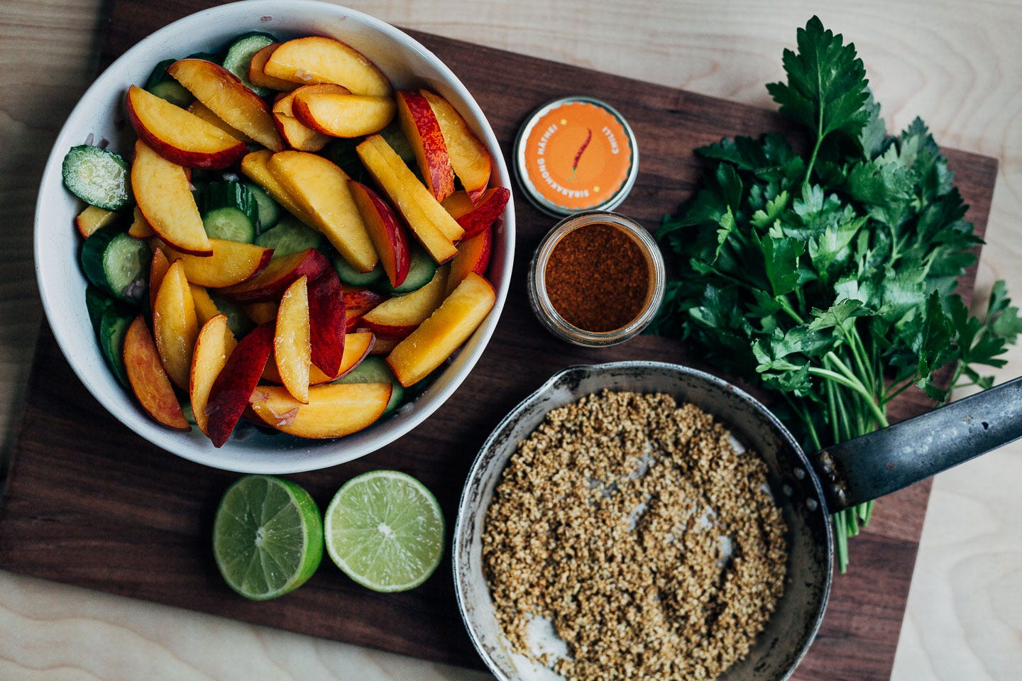 A cutting board with a peach salad, parsley, limes, and a skillet with toasted sesame seeds. 