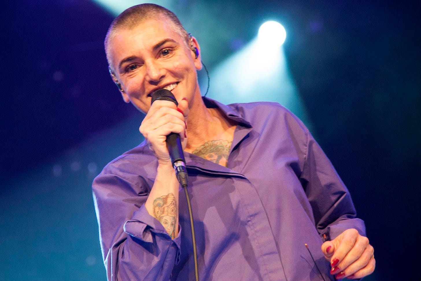 A woman, specifically Sinead O'Connor, wears a purple shirt and sings into a microphone.