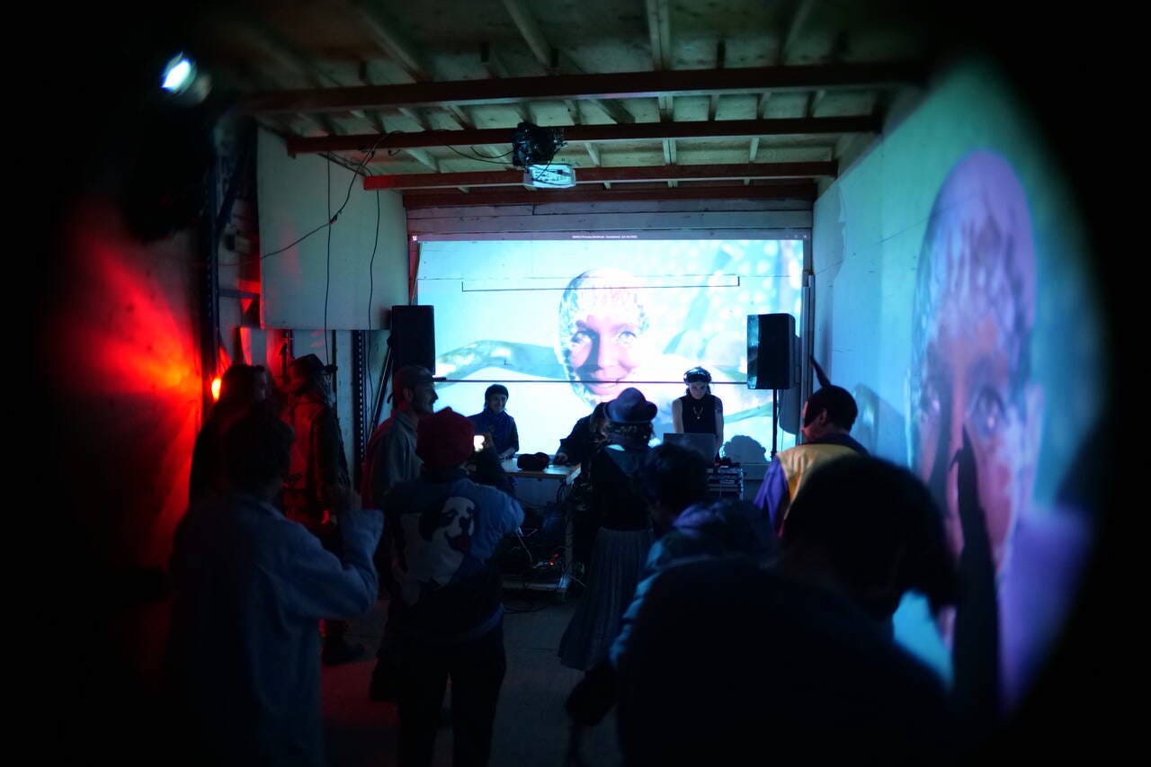 People dancing in a small dark room with dj on the background and two projection of woman face on far and right wall.