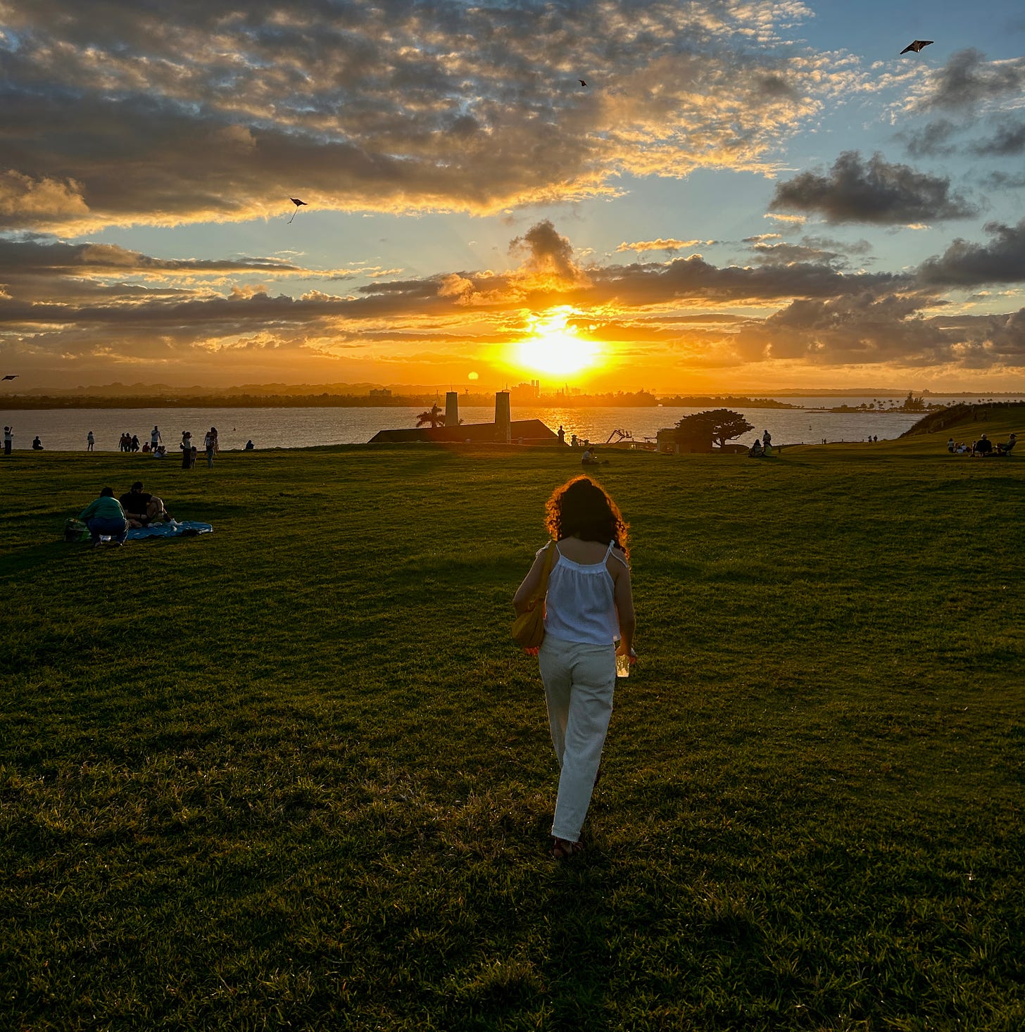 Isa, with dark curly hair and wearing a white blouse and jeans, has her back to the camera. She's walking on a sprawling green lawn toward the sunset, toward the water. Kites are visible floating overhead, and there are other people walking or laying in the park, too.