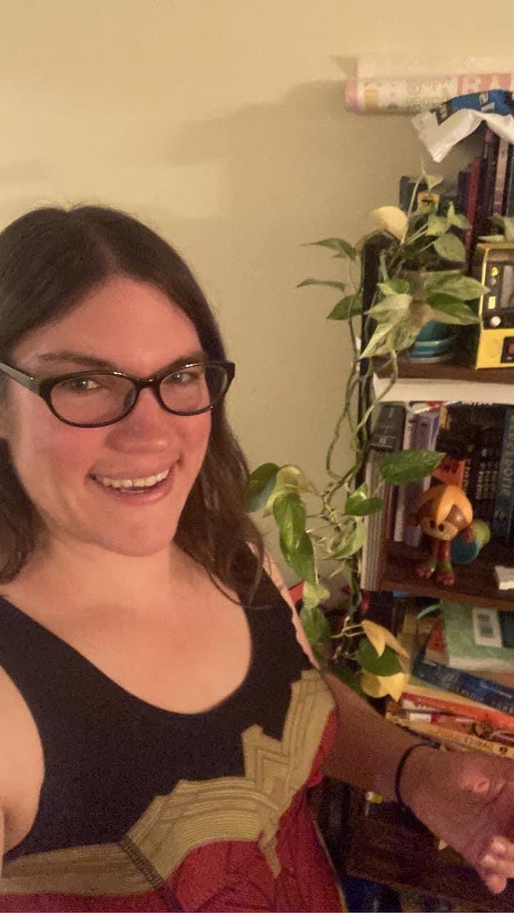 Me (a white woman with brown hair) in a Wonder Woman dress in front of a bookshelf with a plant on it