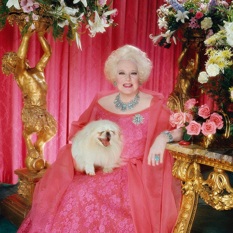 Novelist Barbara Cartland wearing a large pink dress, sitting next to a gold statue, with a small pekinese dog on her lap.