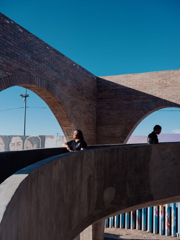 A woman stands facing the sun on a brick and concrete ramp. In the background is the border wall with colorful portraiture and mural painting. 
