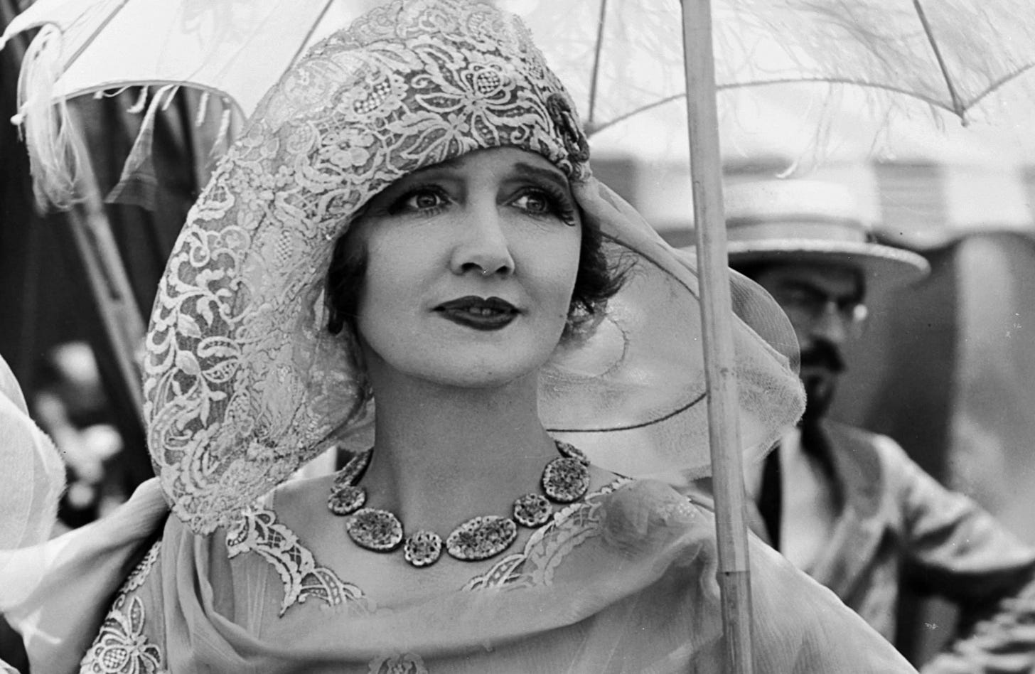 Silent film actress turned Gossip Columnist, Hedda Hopper in a big floppy hat. She is carrying an umbrella, large necklace with each stone as a circle and lots of makeup. 