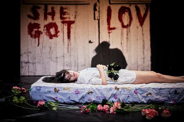 Carolina Bianchi lies unconscious on a mattress, surrounded by flowers, with a shadow looming over her.