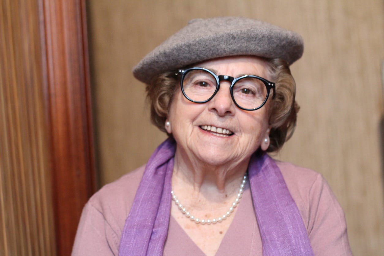 Profile of older white, smiling woman wearing beret-type gray hat on her brown head of hair. She's wearing black, large-rim glasses. large 