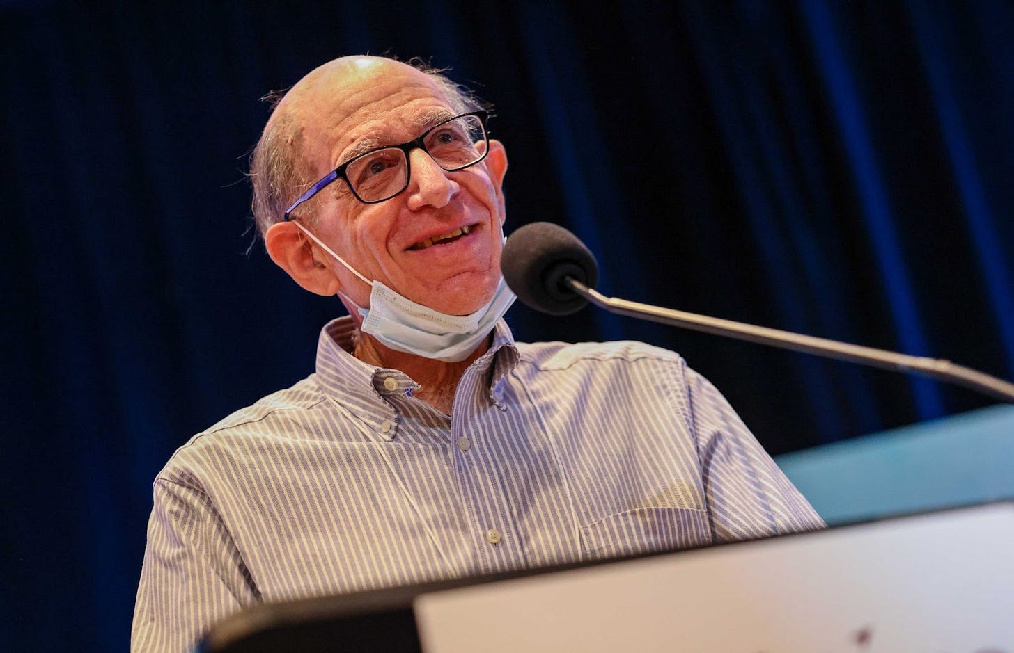 Max talking at a lectern. A older white man with a bald head, blue thin glasses, smiling, with a collard shirt and a covid mask around his chin.