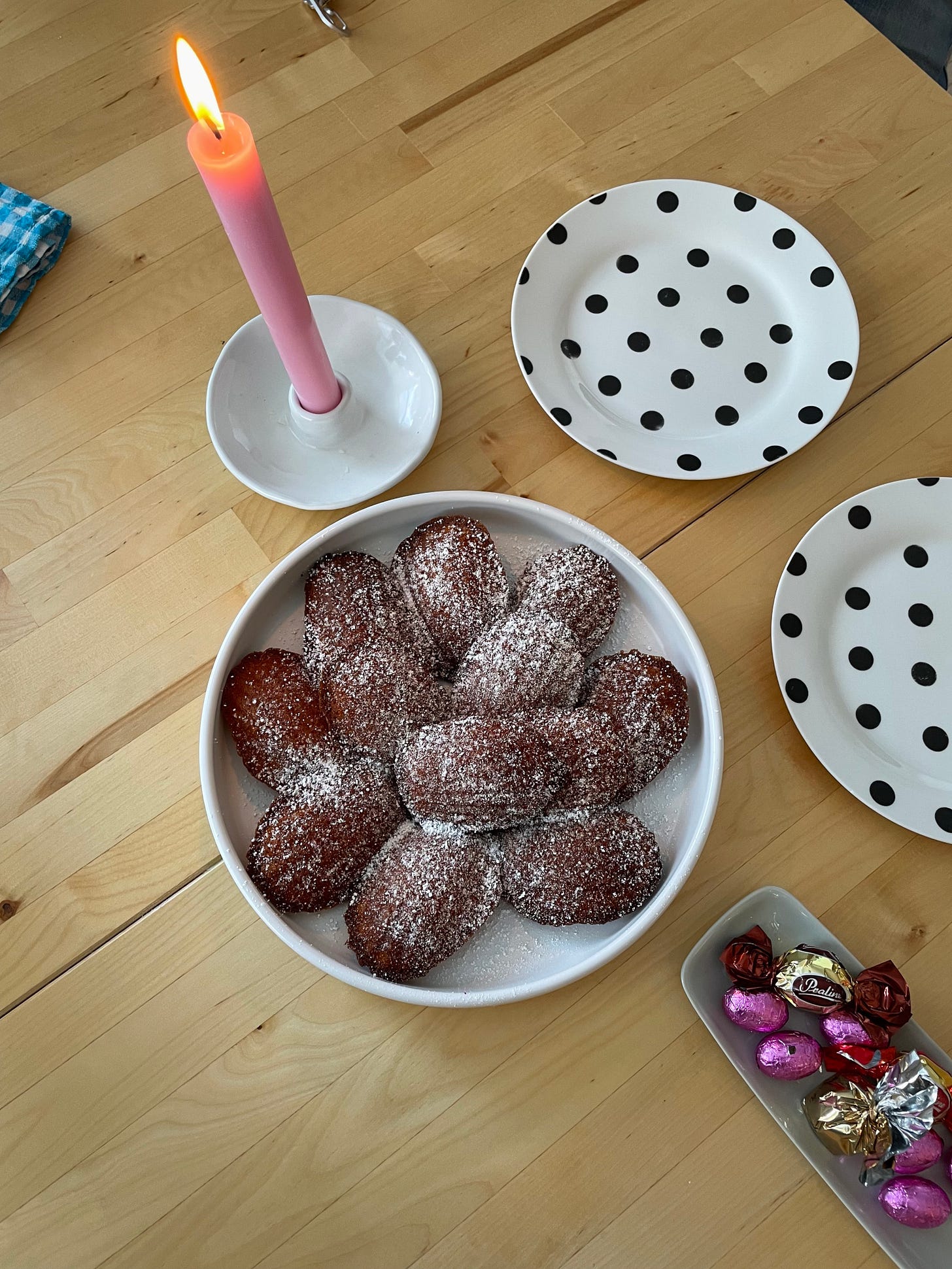 Table setting with a pink candle and a white plate full of madeleine cakes, dusted with icing sugar.