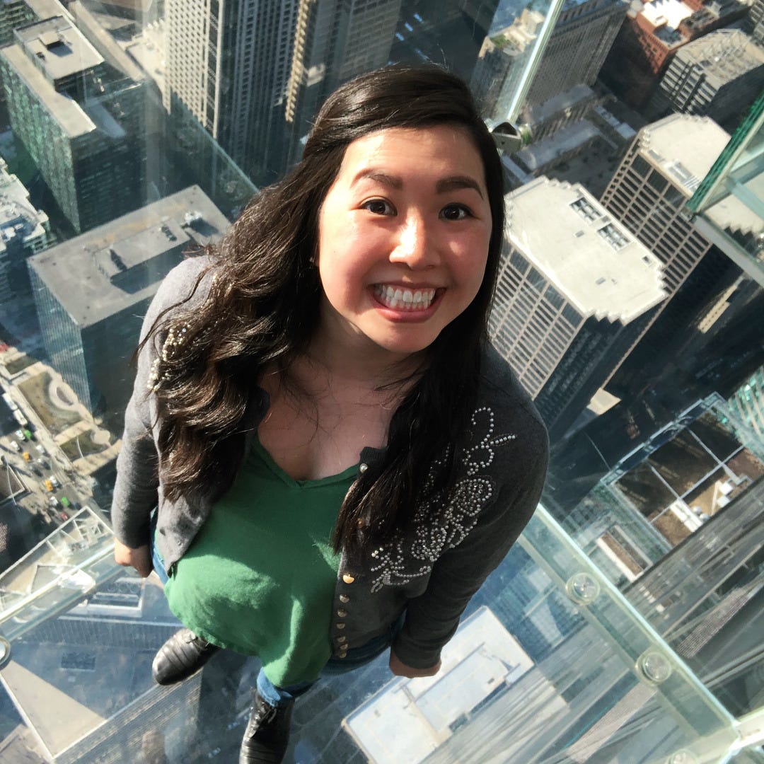 Traci Chee in a green shirt and gray sweater, grinning excitedly while standing in a room of the Willis Tower with a glass floor