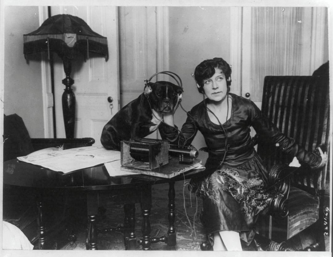 Black and white photo of Mme. Asta Souvrina, “the Bernhardt of Russia,” sitting beside a dog named Buster. Both are wearing headphones, listening to a radiophone device on a table in front of them. Taken around 1922 by Underwood & Underwood. Black and white photo of Mme. Asta Souvrina, “the Bernhardt of Russia,” sitting beside a dog named Buster. Both are wearing headphones, listening to a radiophone device on a table in front of them. Taken around 1922 by Underwood & Underwood.