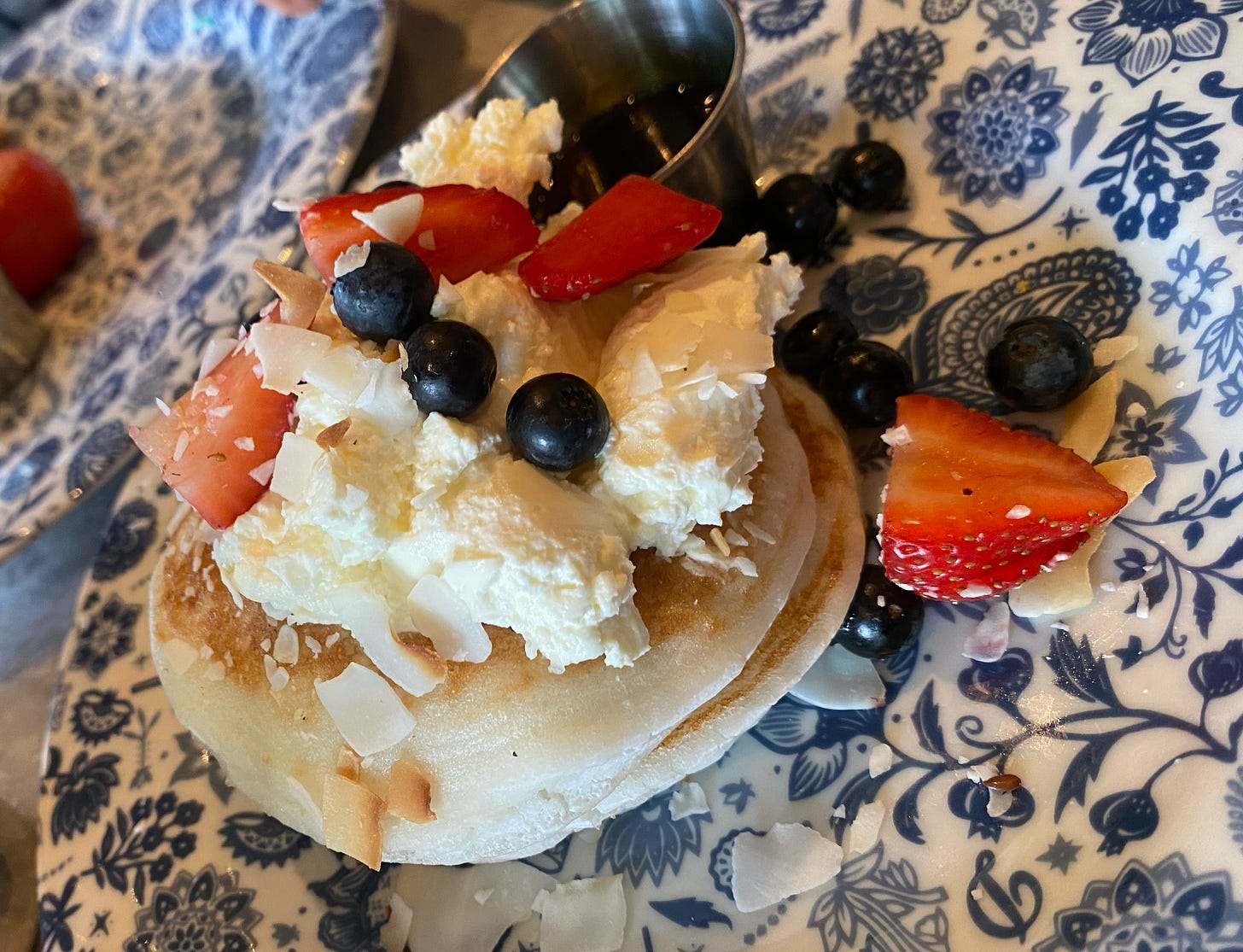 A plate of pancakes topped with blueberries, strawberries, syrup, shrikhand, all on a white porcelain plate with blue designs. There is a metal container for sauce.