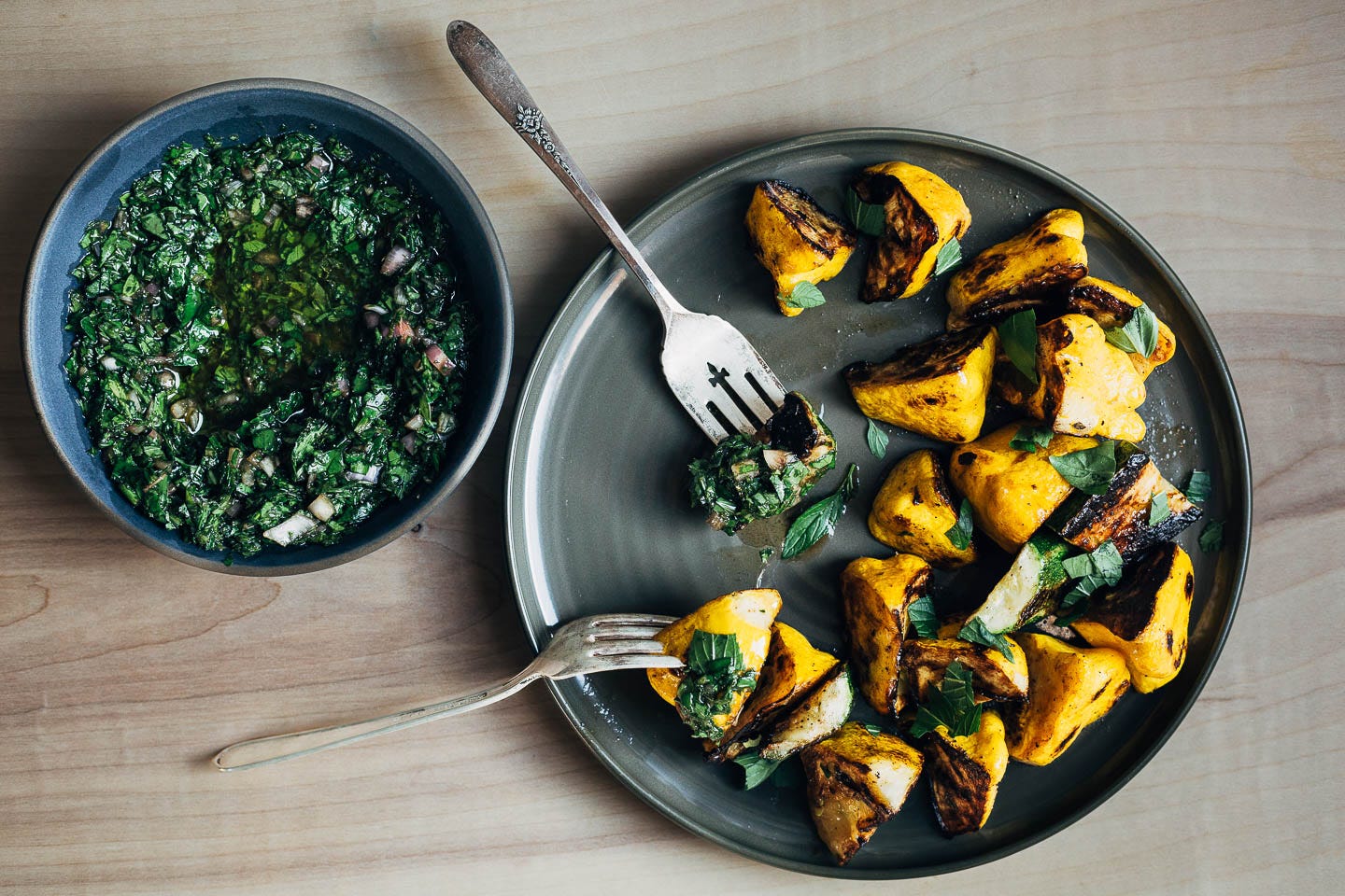 A small bowl of chimichurri alongside a plate with seared squash and fresh herbs. 