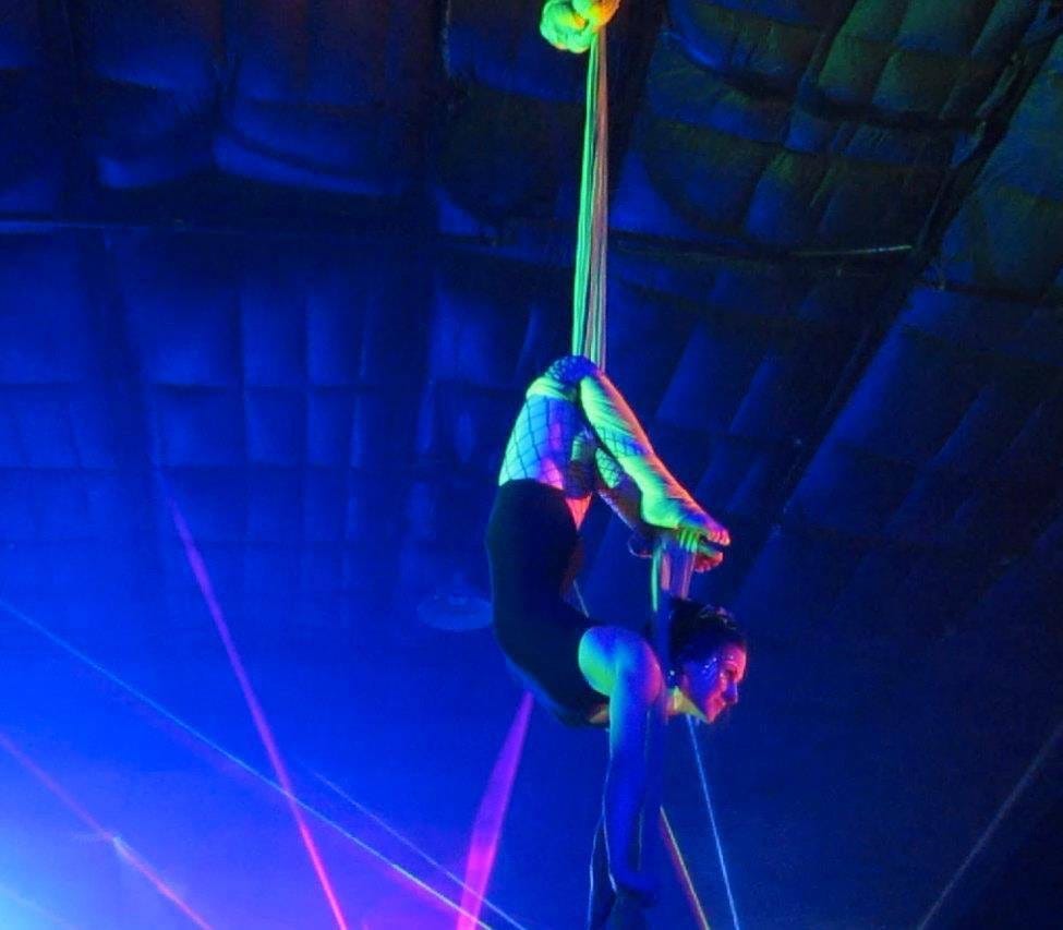 Lyric, in a dark room lit with black-lights and lasers, is hanging from the rafters in a building from their knees on neon-green aerial silks, in a pretzel shape, with their feet behind their head.