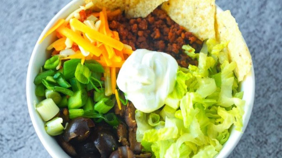 A bowl of taco salad featuring layers of lettuce, sour cream, green onions, carrots, seasoned ground meat, and olives, garnished with a tortilla chip.