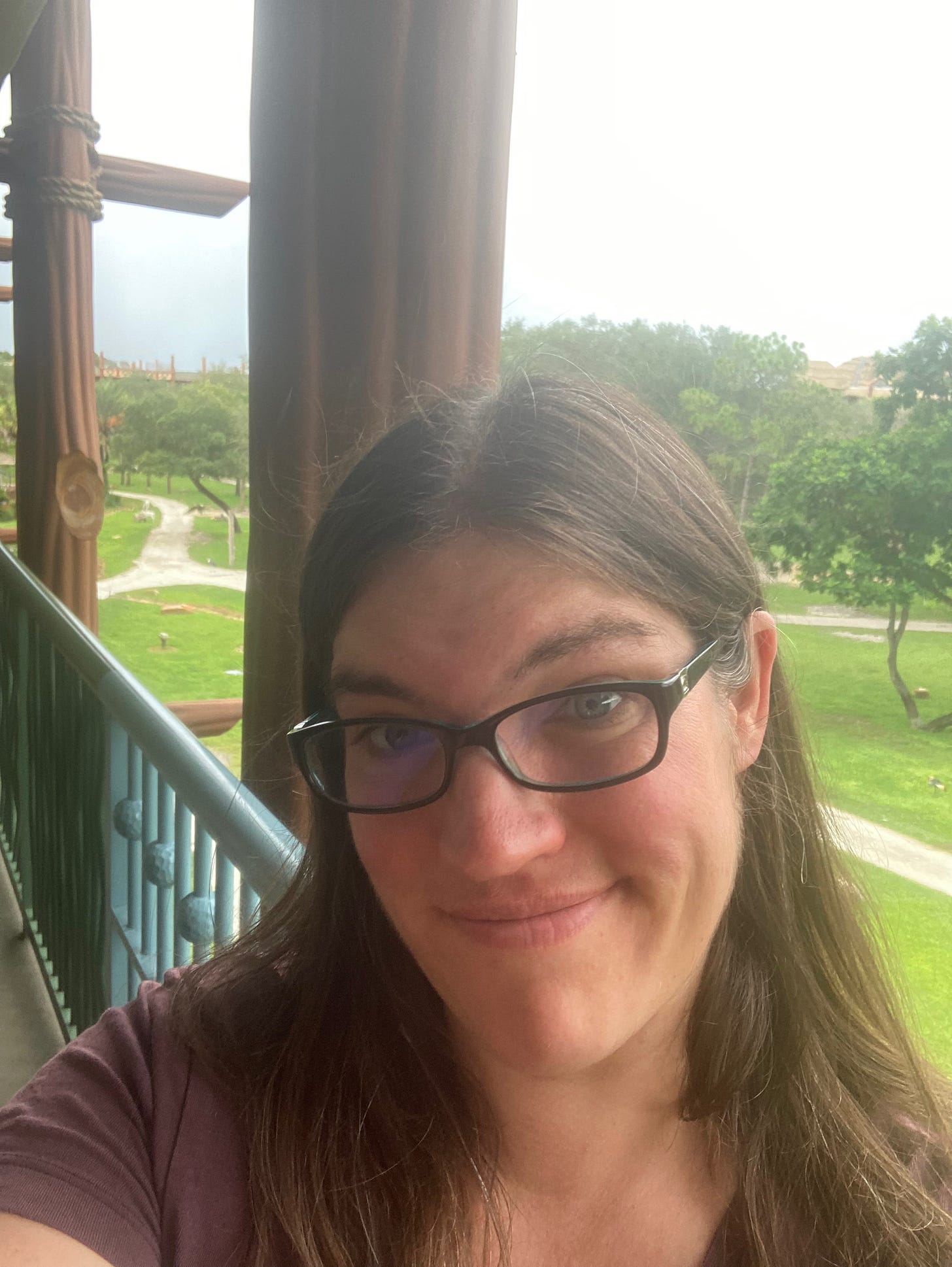 Me (a white woman in a burgendy t-shirt) looking at the camera with a skeptical look while standing on the balcony of a hotel with grass and trees below