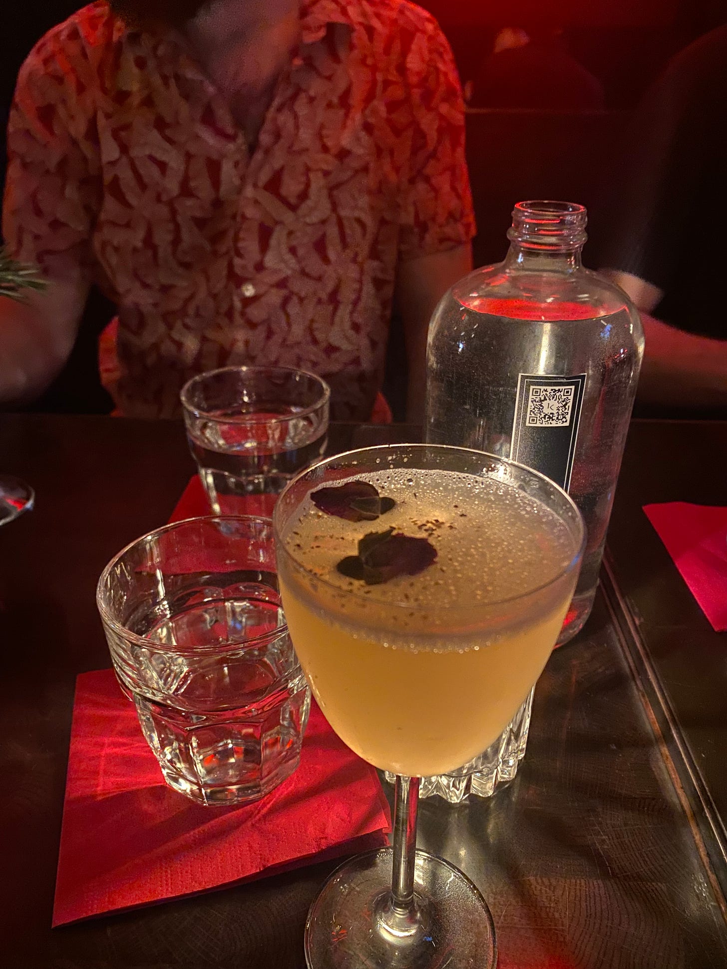 my Skeleton's Kiss cocktail, with two dried rosebuds and crushed chili floating on top. Jeff, wearing a red and white patterned shirt, is visible across the table.