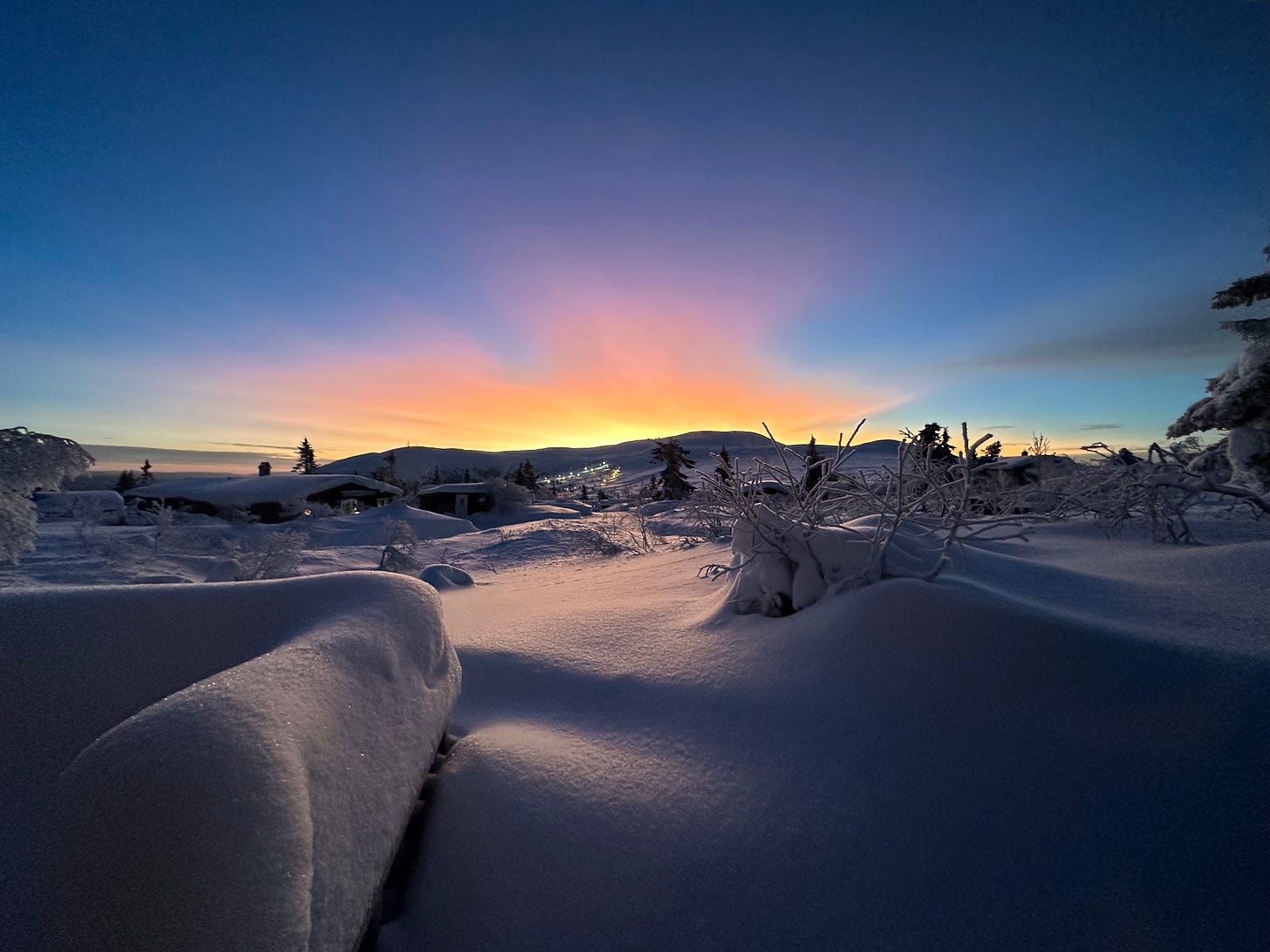 Cenário coberto de neve alta com uma montanha ao fundo e o sol nascendo atrás dela.