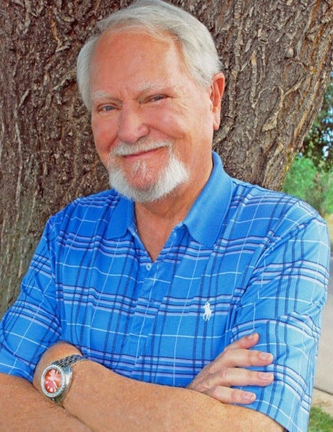 Clive Cussler with white hair and goatee in a blue shirt with arms crossed smiling