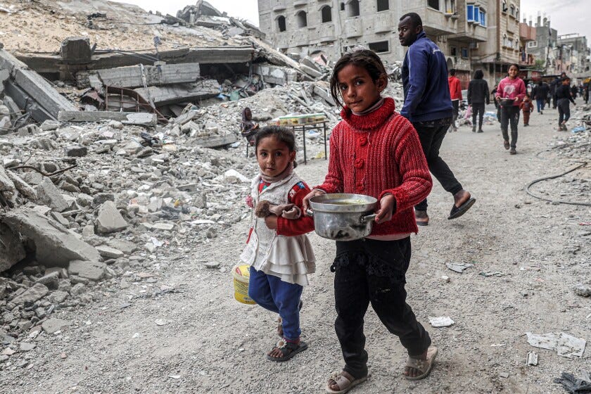 Two kids wearing sweaters and sandals walk past the rubble of a collapsed building, one carrying a pot of food.