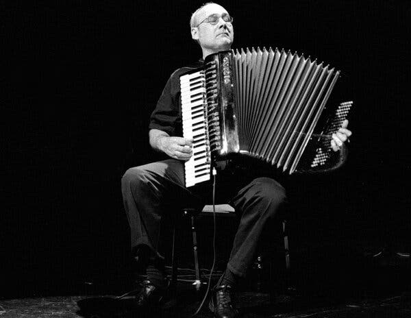 A black-and-white photo of Guy Klucevsek, a bald man wearing glasses and a short-sleeved shirt, seated on a chair playing an accordion. His eyes are closed.
