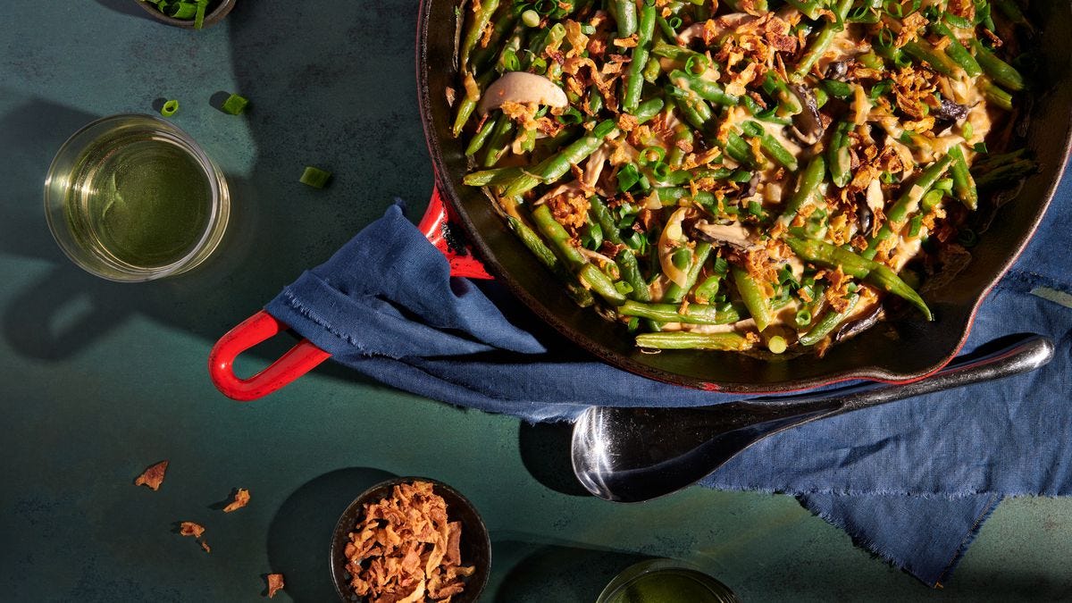 Green bean casserole in a red castiron skillet. Next to the skillet are two drinking glasses, small bowls of sliced scallions and fried shallots. A blue napkin is draped over the skillet handle.