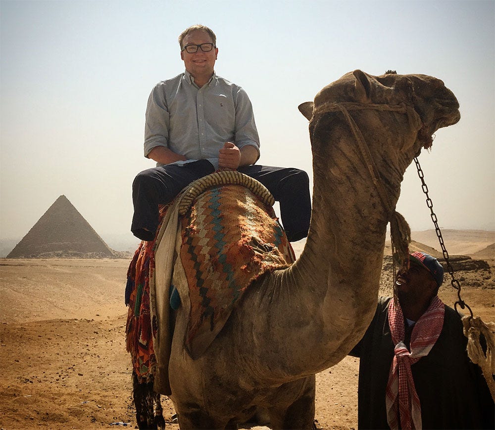 John seated on a camel in front of an Egyptian pyramid.