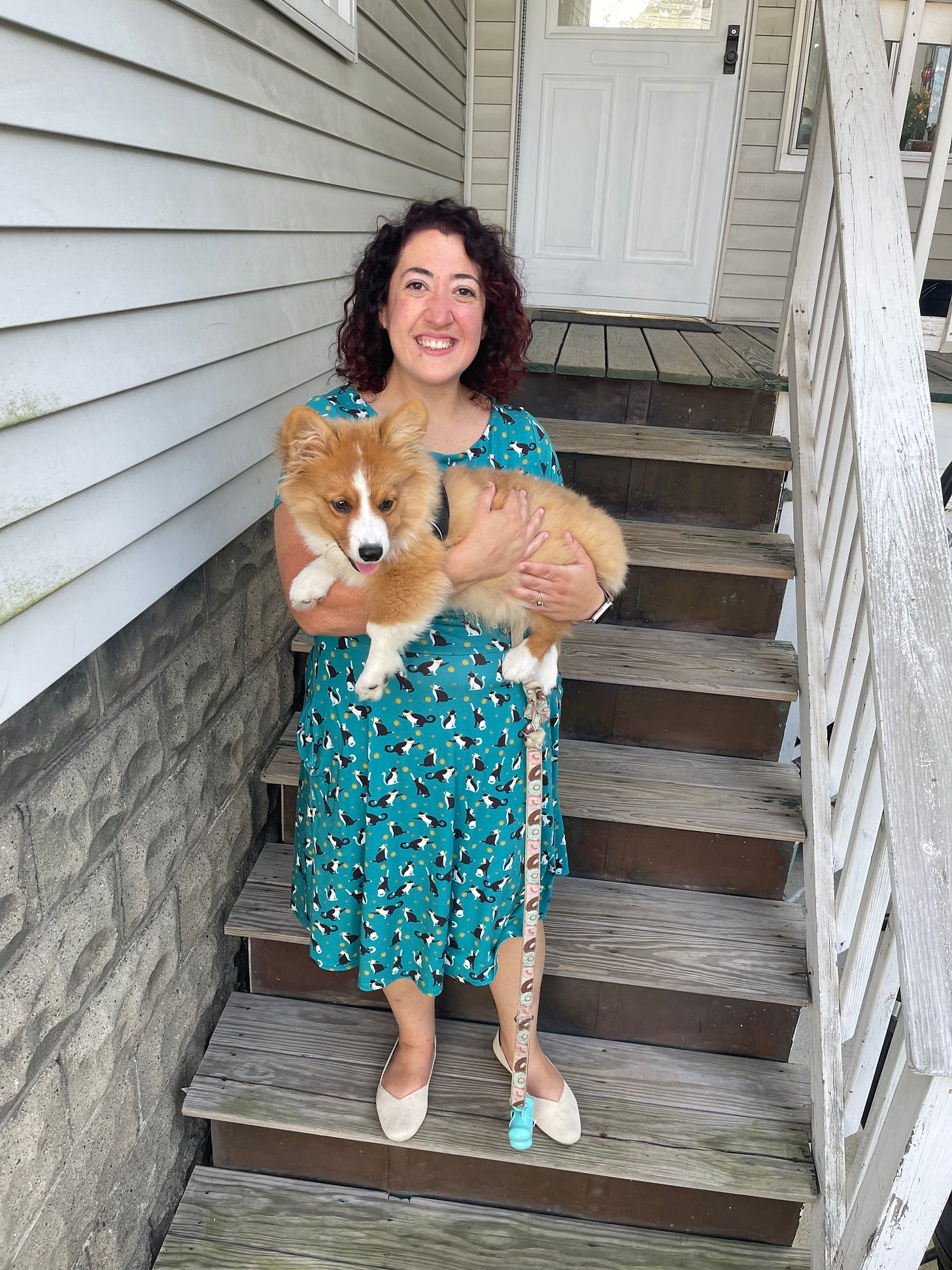 A woman in a blue patterned dress stands smiling on the stairs while holding a corgi puppy in her arms. 