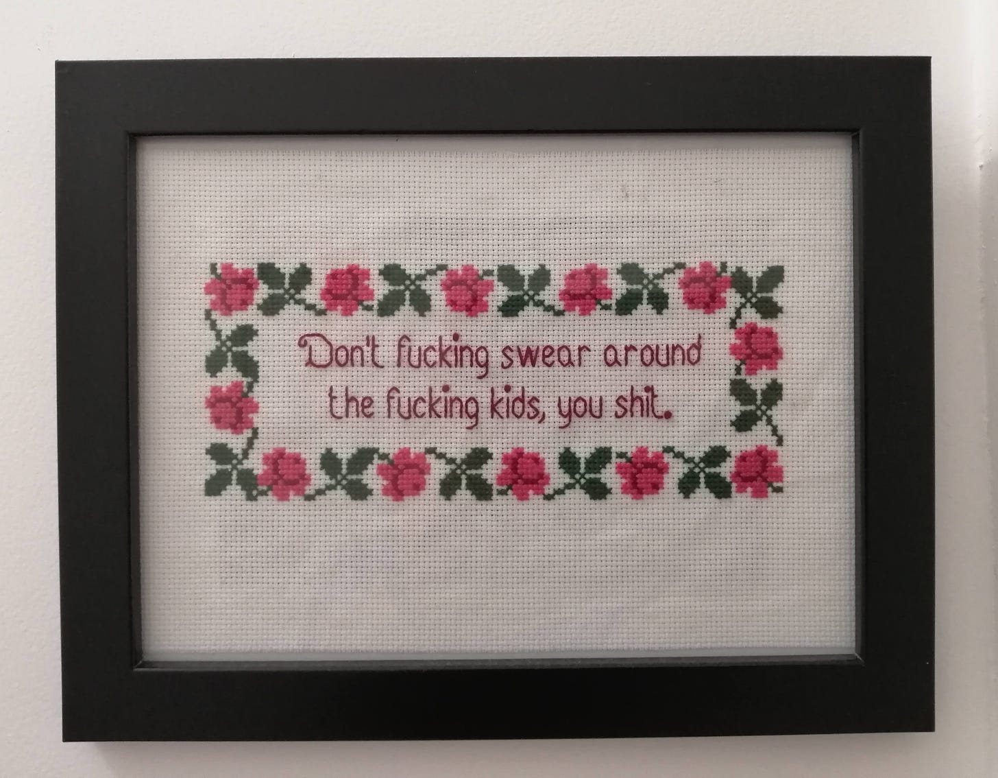 Completed cross stitch in a frame with a cute floral border. The text reads " don't fucking swear around the fucking kids, you shit"