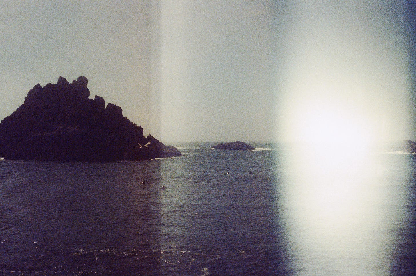 A slightly grainy film photograph of the ocean, with a large rock formation off to the left, and a thick light leak on the right third of the frame.