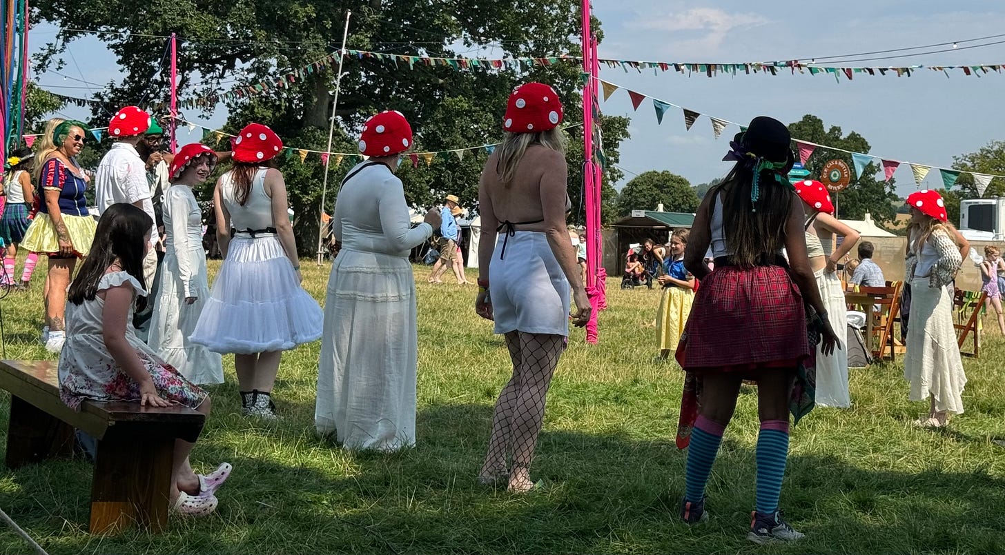 A grassy field with trees in the background.  A group of woman wearing white are all adorning red mushroom hats with white spots.  Could be a toadstool?  They are getting ready to be taught a folk dance.