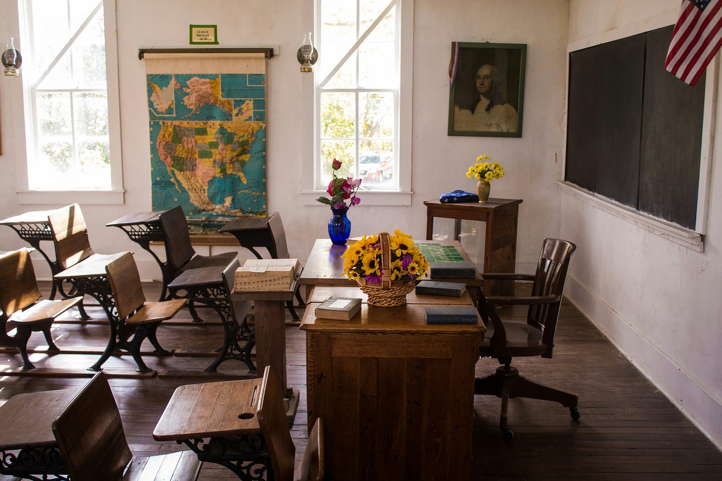 A color picture of an empty, antiquated classroom with wooden desks, a map of the U.S., a U.S. flag, and a picture of George Washington.