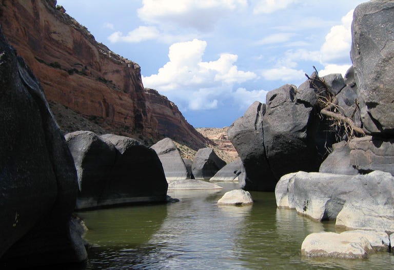 View from the surface of a rippling green river in a narrow passageway between huge gray rocks, bordered by red canyon walls