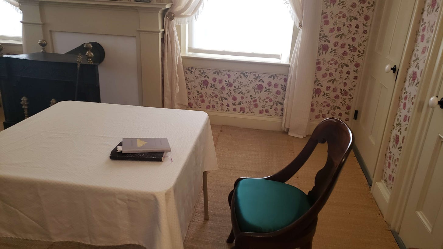 A card table with a white tablecloth and a period parlor chair with a deep green silk cushion. Set up in a corner of Emily Dickinson's bedroom by the fireplace and the window. 