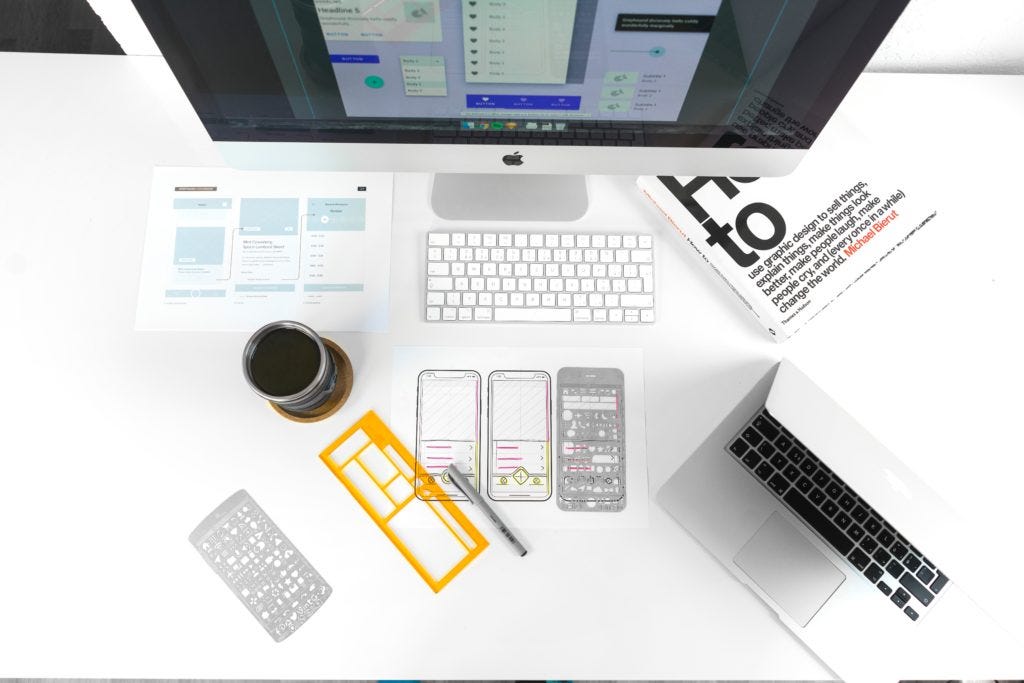 desk of a designer. a desktop computer, a laptop, coffee, a design book, and prototypes