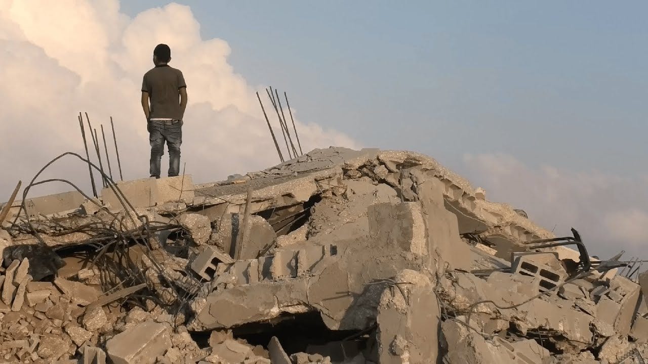 A Palestinian man standing on a pile of rubble looking away from the camera.