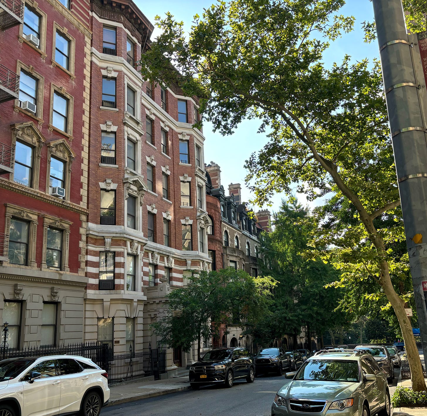 A red brick building with two sets of bay windows on each floor on a quiet tree-lined street full of townhouses. Riverside Park is visible at the end of the street. There is a small plaque on the ground floor that can't be read from this distance.