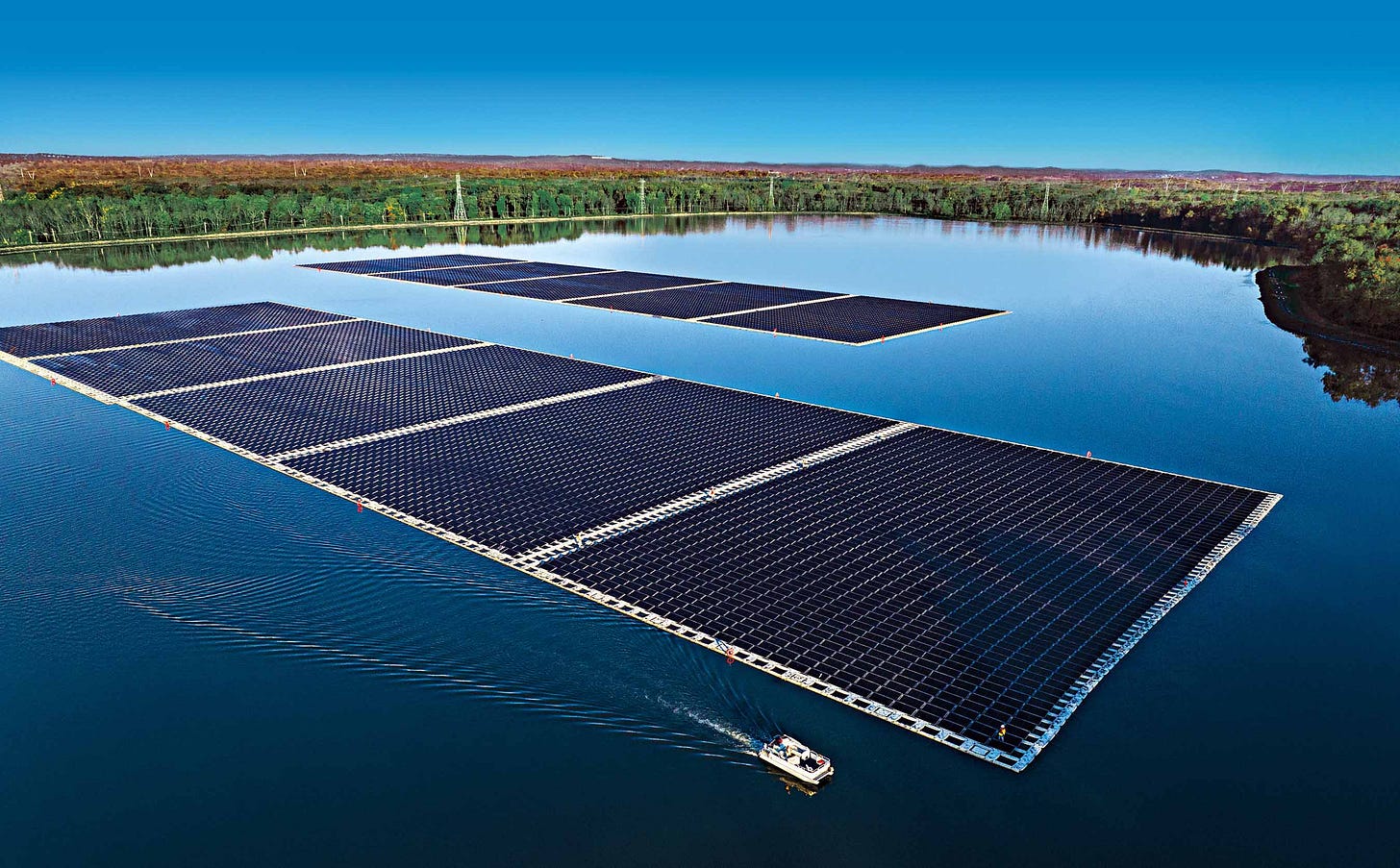 Floating solar panels on a large body of water with a small boat nearby; forested shorelines in the background under a clear blue sky.