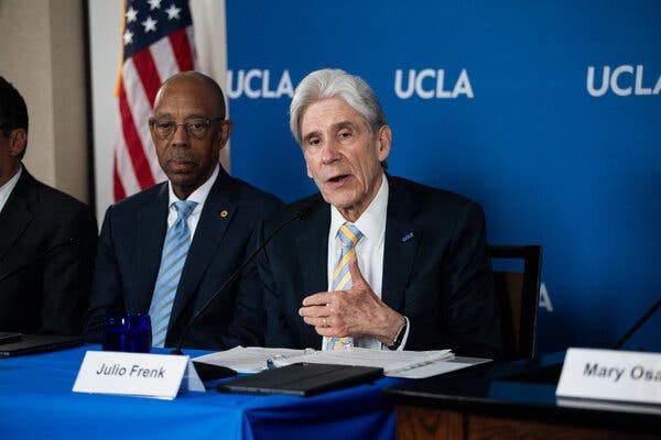 Julio Frenk, wearing a blue jacket and blue and gold striped tie, speaks at a news conference table. To the left is Michael Drake, the University of California president, wearing a blue jacket and blue and gray striped tie. A U.C.L.A. blue backdrop is behind them.