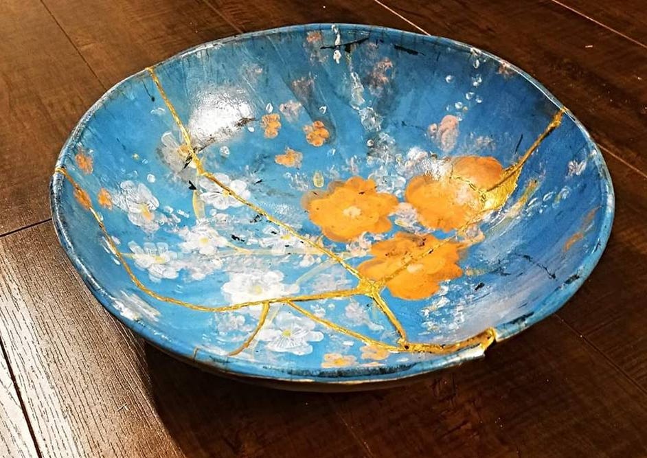 A blue bowl with orange flowers, cracked in several places but glued back together with gold; the bowl is on a wooden table