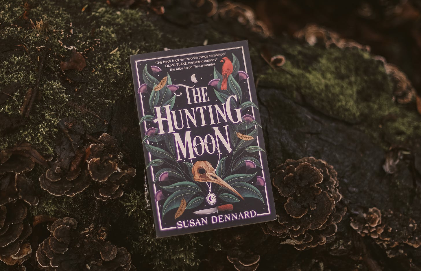 A photograph of the UK edition of The Hunting Moon with the book resting atop a mossy log