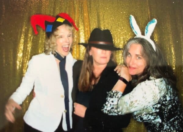 From left: E. Jean Carroll in a costume jester hat, white blouse and black tie; Mary L. Trump in a black felt hat pulled low over her eyes, wearing a black top and a black jacket over it; and Jennifer Taub, who has placed her hands on Trump’s shoulder and mugs for the camera wearing a headband with fabric bunny ears. They’re against a glittery, gold backdrop and the photograph is somewhat out of focus; it looks like a party shot.