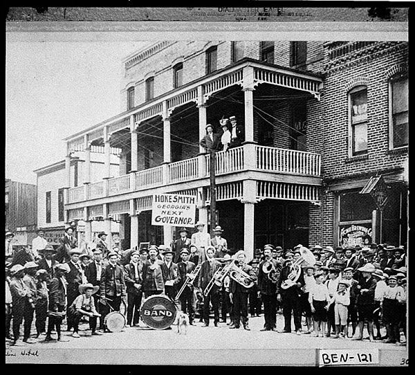 People standing in front of two-story building with band, and sign reading "Hoke Smith. Georgia's First Governor"