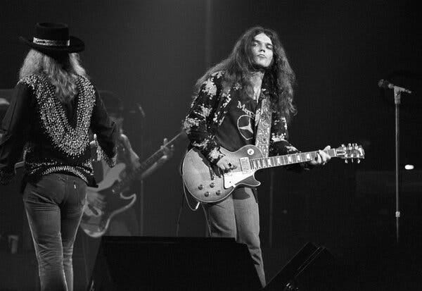 A black-and-white photo of Gary Rossington playing electric guitar. He has long brown hair, parted in the middle, wearing jeans and a long-sleeve print shirt.
