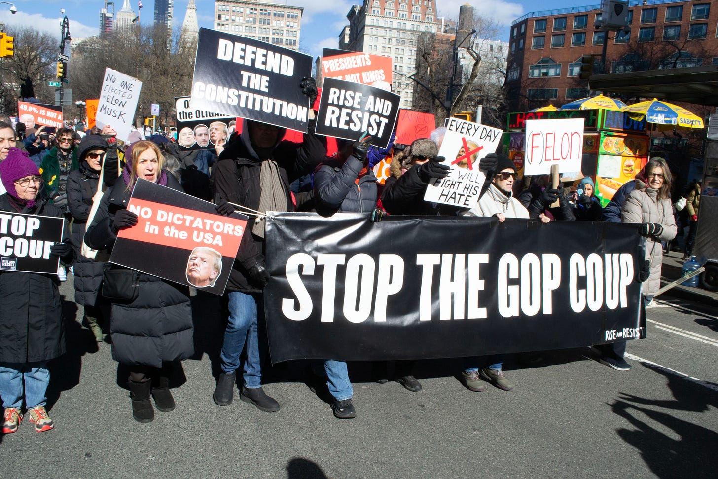 A group of protesters hold signs that say, from left to right "No one elected Elon Musk," "No dictators in the USA," "Defend the Constitution," "Rise and Resist," "Every day we will fight this hate," and "(F)ELON," with a larger sign held up by multiple protesters saying "Stop the coup" A group of protesters hold signs that say, from left to right "No one elected Elon Musk," "No dictators in the USA," "Defend the Constitution," "Rise and Resist," "Every day we will fight this hate," and "(F)ELON," with a larger sign held up by multiple protesters saying "Stop the coup"