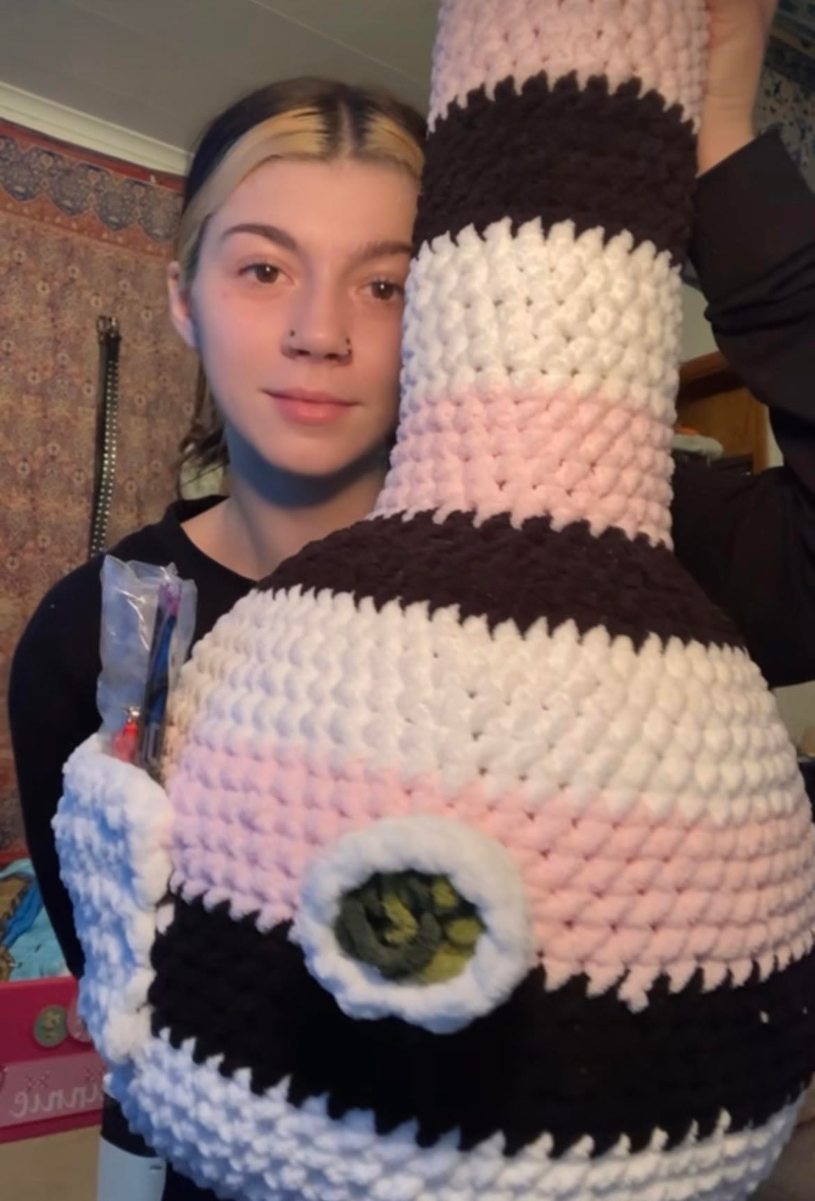 Women holding large crocheted bong plushie, colors are pink, white, and black.